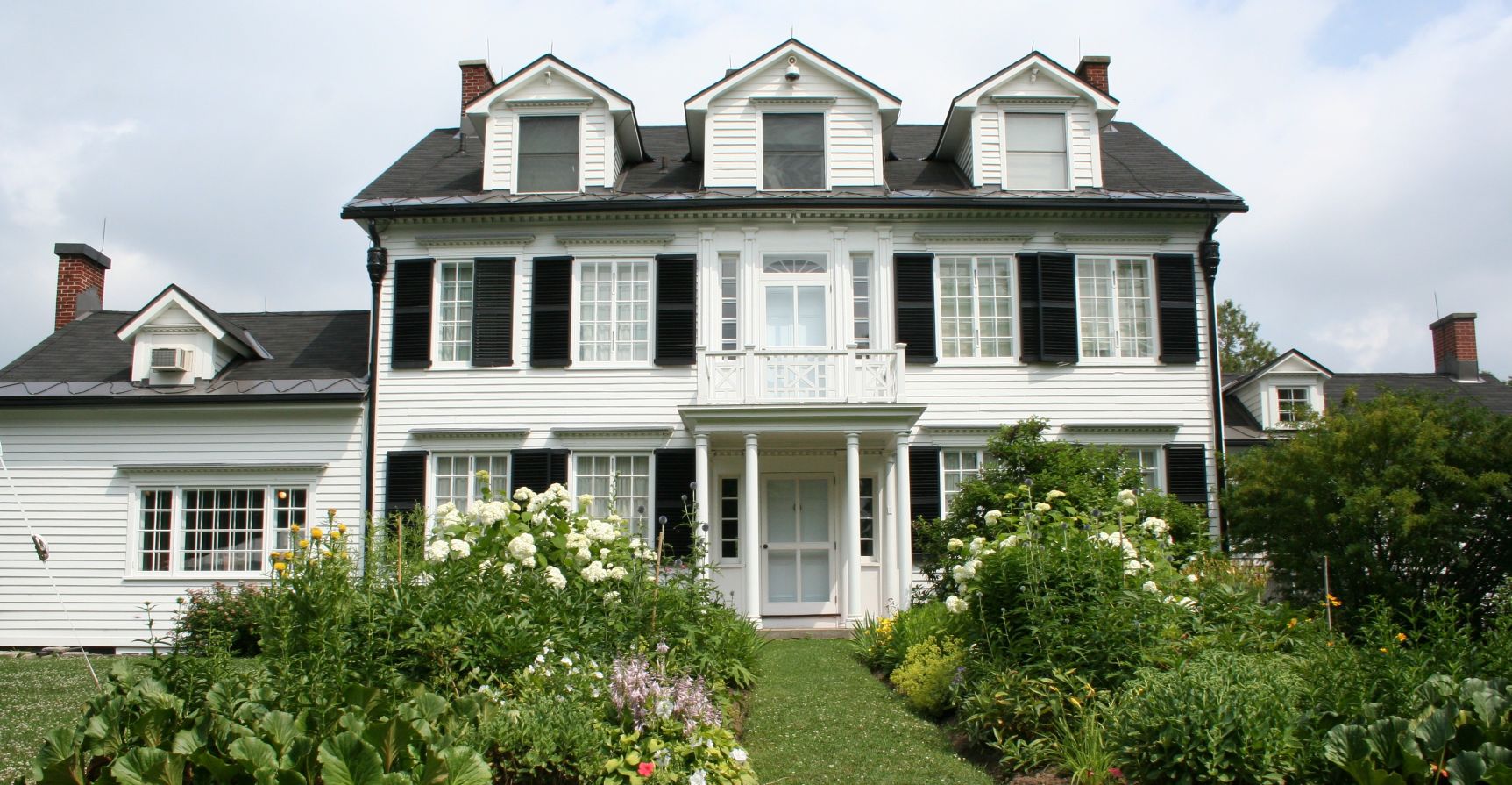 Exterior view of Ottawa's oldest wood-frame house. It is white with black shutters, features a second storey balcony, and lush summer gardens.